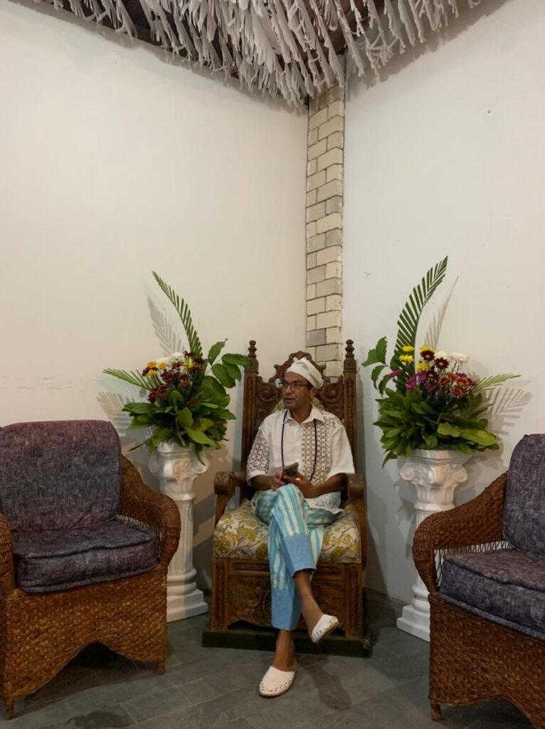 Pai Adauto é o guia espiritual dos fiéis que frequentam o seu terreiro. Na foto, ele está sentado de pernas cruzadas em uma poltrona do terreiro. Ele veste chapeu, sapatos e camisa de mangas curtas brancos e uma calça azul clara. Ele comenta sobre a intolerância religiosa sobre as religiões de matriz africana.